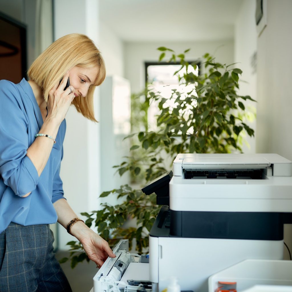 Smiling businesswoman printing documents while talking mobile phone in the office.